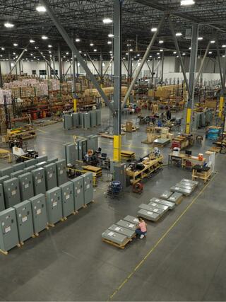 A spacious warehouse filled with organized shelves, machinery, and goods, featuring workers and various equipment throughout the area.